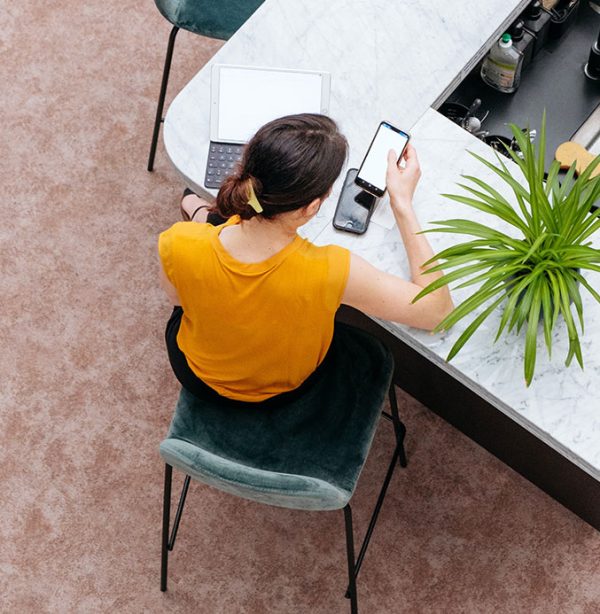 A woman in a yellow top sits at a marble counter using a smartphone, with a laptop and a green plant nearby.