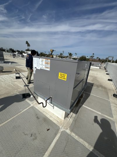 A large HVAC unit is installed on a rooftop, with warning labels visible. Two people are nearby, and shadows are cast on the ground. The sky is partly cloudy.