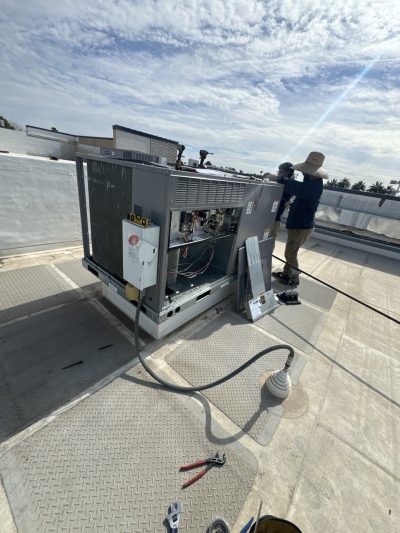 A technician in protective clothing works on an open rooftop HVAC unit under a partly cloudy sky, with tools and cables visible nearby.