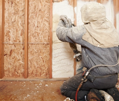 A worker in protective clothing sprays foam insulation between wooden wall studs in a building under construction.