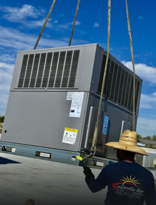 A worker wearing a straw hat guides a large HVAC unit being lifted by straps onto a rooftop under a partly cloudy sky.
