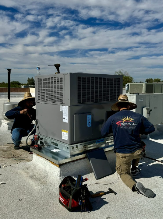 Two technicians in hats are servicing a rooftop HVAC unit under a partly cloudy sky, with tools and equipment placed nearby.